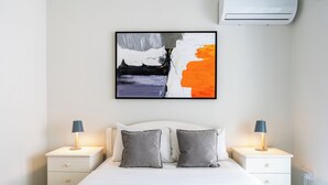 A modern bedroom with a large bed, two bedside tables, a wall-mounted air conditioner, and a framed abstract painting. - Sweet Retreat 2-BR Apt. in Mackenzie (Larnaca)