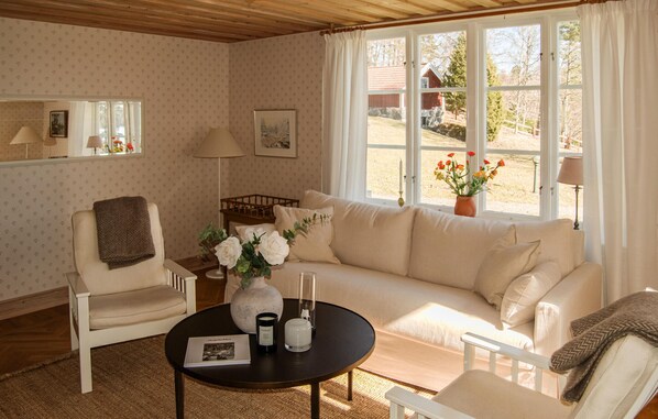 Ein gemütliches Wohnzimmer mit einer Couch, Sesseln, einem Couchtisch und Blick auf einen roten Bauernhof durch große Fenster.