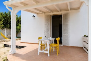 Un porxo cobert amb una taula blanca i dues cadires grogues, una paret blanca i una porta que dona a l'interior.