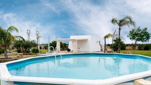 Una piscina con solarium bianco, un'area salotto coperta e palme sullo sfondo.