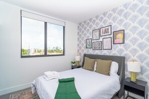 A neatly made bed with white bedding, a green towel, and a city view through a large window. - Miami City Escape 2BR Suite Near Brickell & Miami Beach (Miami)