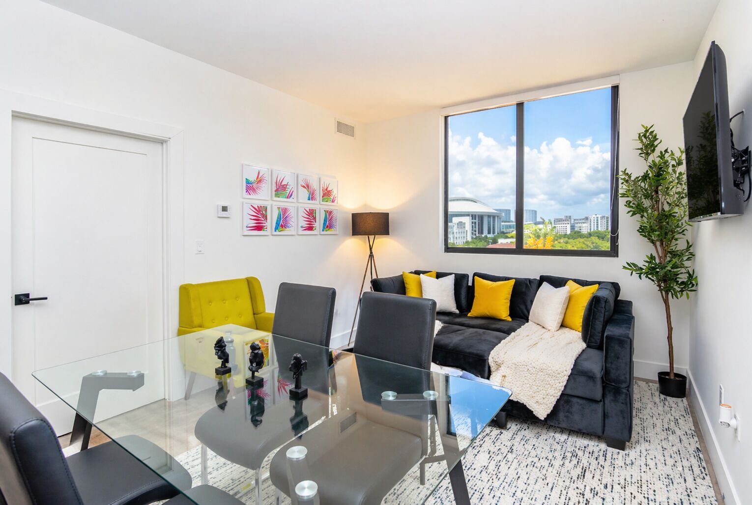 A modern living room with a glass table, black and yellow furniture, a large window with a city view, and framed artwork on the wall.