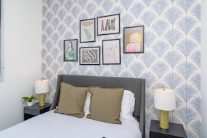 A neatly arranged bedroom with a patterned wallpaper, a bed with two pillows, a bedside table with a lamp, and framed artwork on the wall.