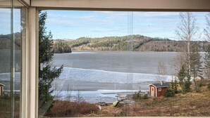 En vy genom ett fönster som visar en sjö med en träbrygga, en röd byggnad och omgivande träd.