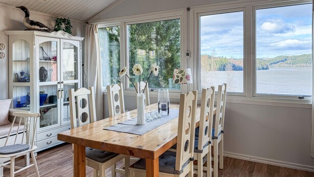En matplats med ett träbord och vita stolar, ett skåp med glasdörrar och utsikt över en sjö genom stora fönster.