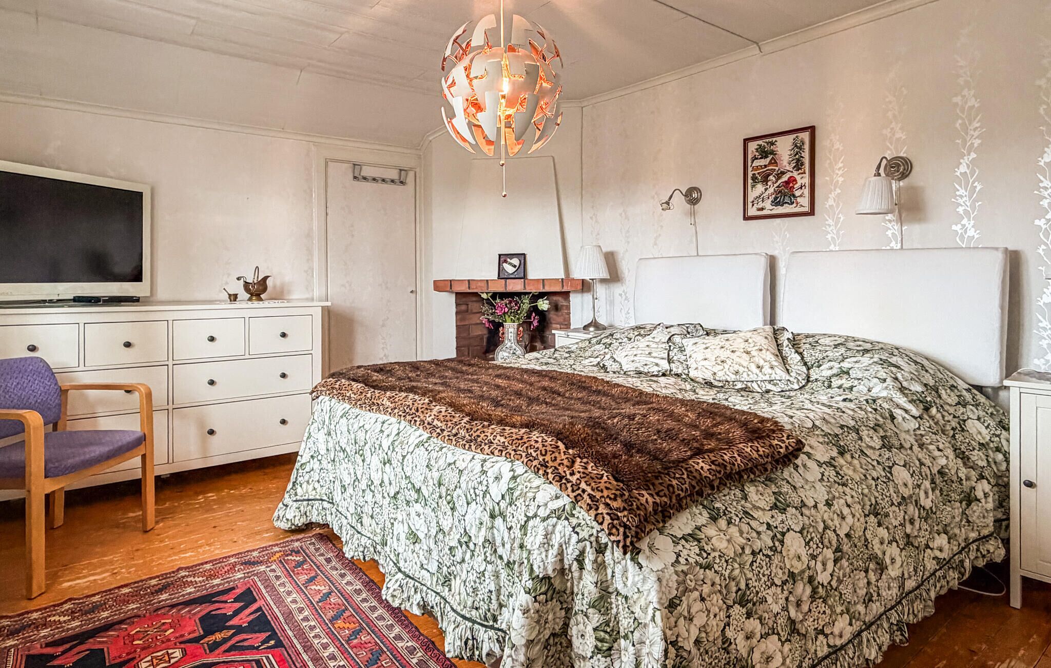 Un dormitorio con cama, televisor, cómoda, sillón, chimenea y lámpara colgante.