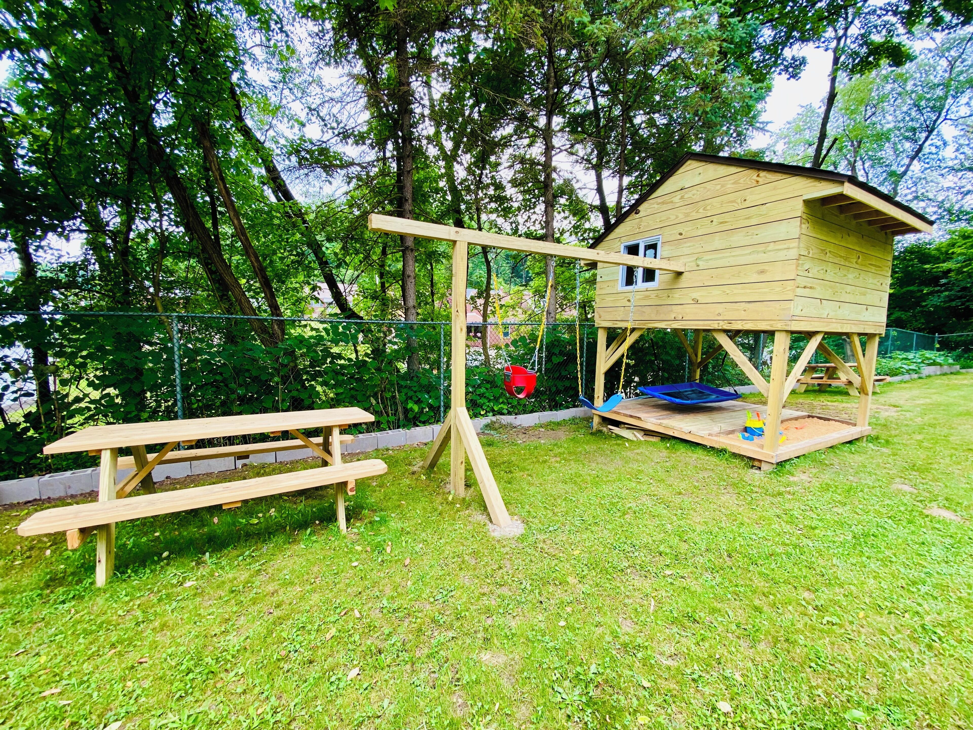 A wooden playhouse with a swing set and a picnic table in a grassy yard.