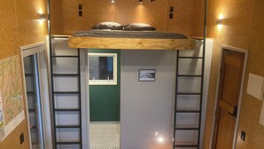 Een kleine kamer met een hoogslaper, inbouwplanken, een klein raam en een deur naar een badkamer.