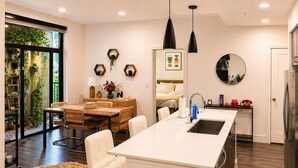 A modern kitchen with a central island, a dining area with a wooden table and chairs, and a view of a bedroom through an open door. - NRG Jungle Highrise - Balcony & Pool View - Walk to FIFA/NRG/Med Center (Houston)