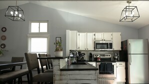 Una cocina con armarios blancos, microondas, estufa y refrigerador. Hay una zona de comedor con sillas y una mesa.