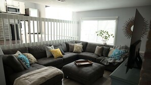 Una sala de estar con un sofá seccional, un reposapiés y una planta en maceta junto a una ventana.