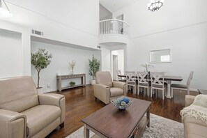 A spacious living room with a high ceiling, a dining area with a white table and chairs, and a balcony with a railing. - Oxnard Summer Escape with Pool + Hot Tub (Oxnard)