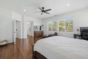 A bedroom with a large bed, a ceiling fan, a dresser, a TV, and a window with blinds.