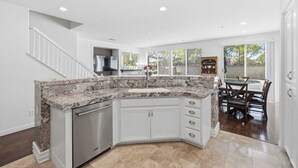 A kitchen with a granite countertop, white cabinets, a dishwasher, and a dining area with a table and chairs. - Oxnard Summer Escape with Pool + Hot Tub (Oxnard)