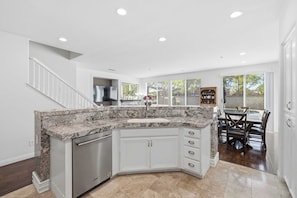 A kitchen with a granite countertop, white cabinets, a dishwasher, and a dining area with a table and chairs. - Oxnard Summer Escape with Pool + Hot Tub (Oxnard)