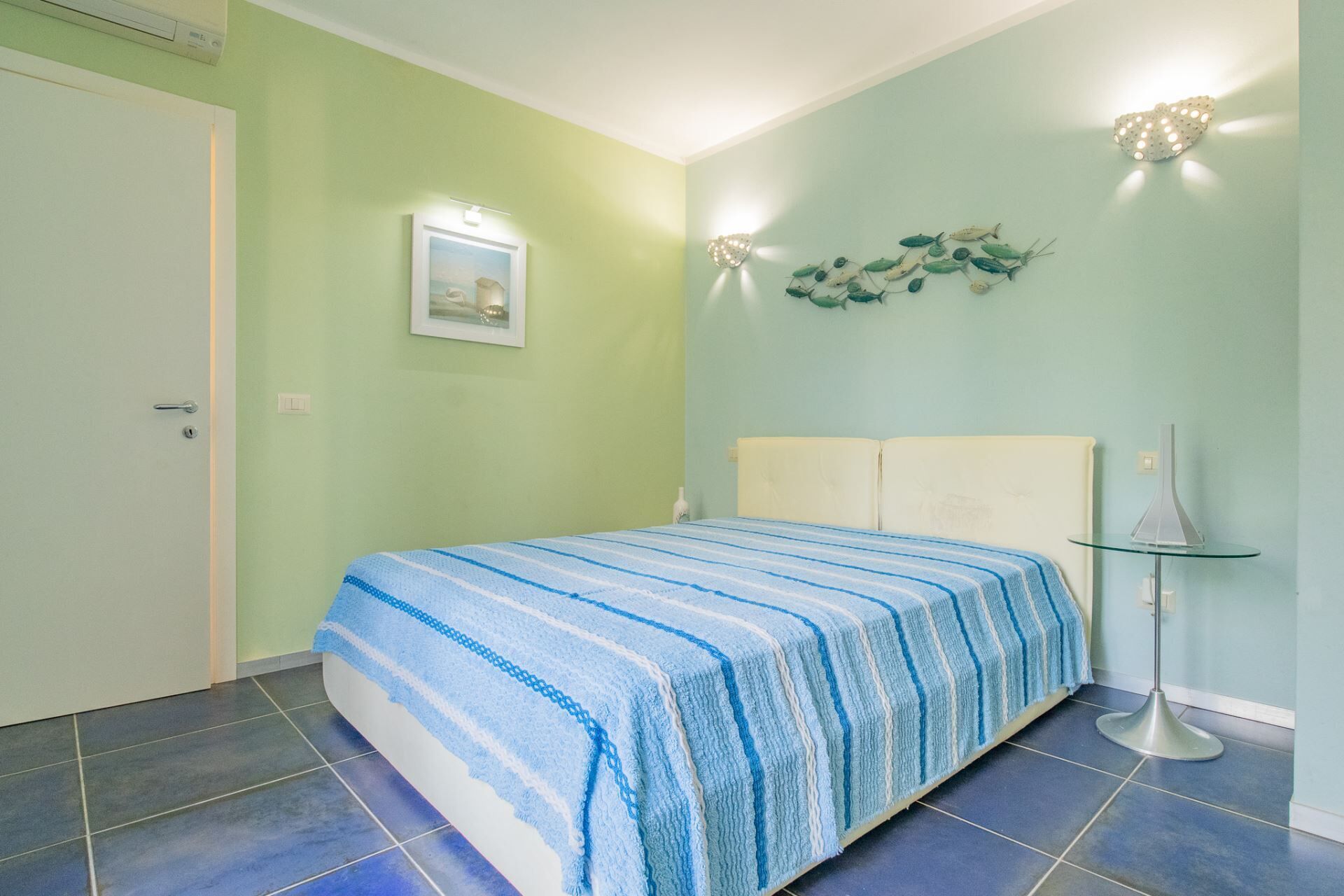 Un dormitorio con cama, mesita de noche, lámpara de pared e imagen enmarcada.