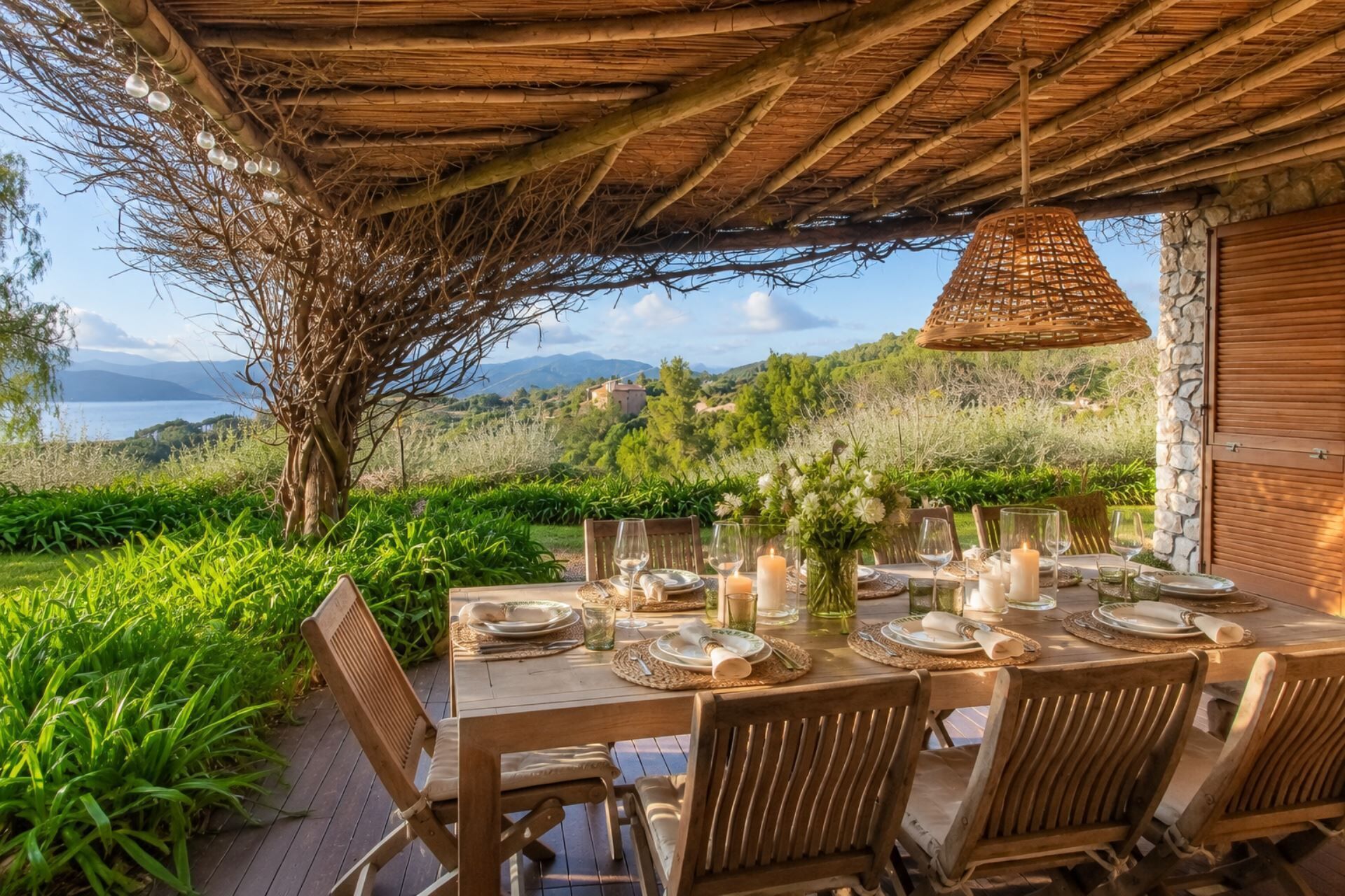 An outdoor dining area with a table set for six, surrounded by lush greenery and overlooking a scenic landscape.