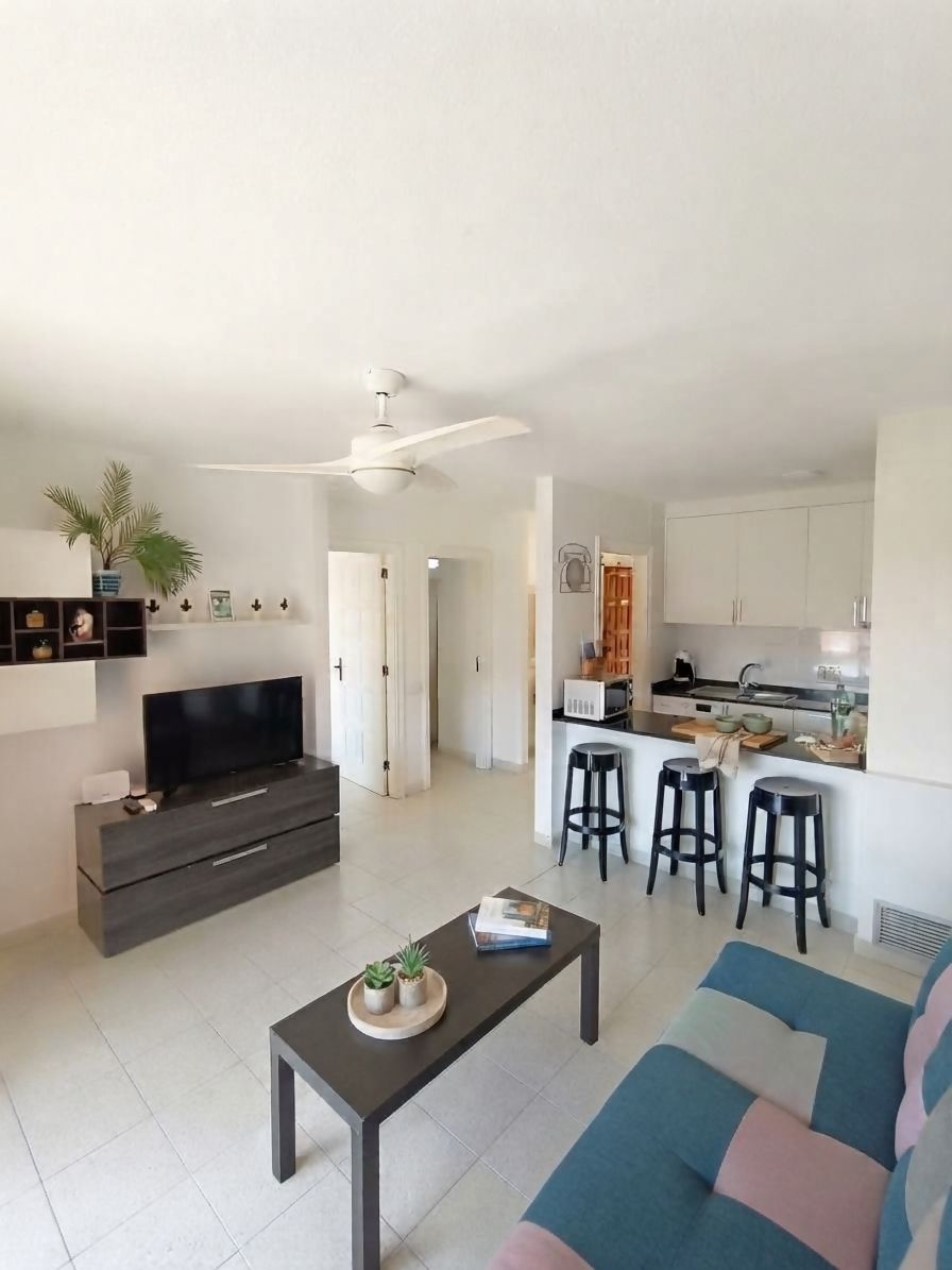 A modern living room with a flat-screen TV, a dark sofa, a coffee table, and a kitchen area with a bar stool.