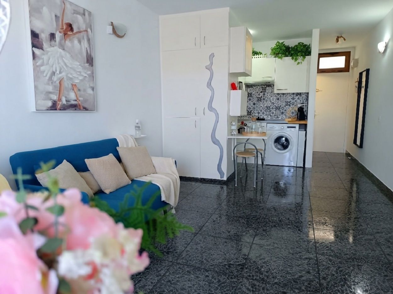 A living room with a blue sofa, a white wall with a painting, a kitchen area with a washing machine, and a tiled floor.