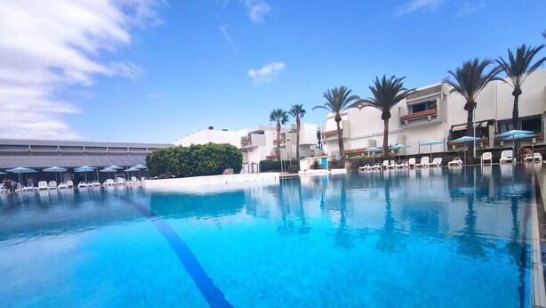 A swimming pool with lounge chairs and umbrellas, surrounded by palm trees and buildings. - RenTenerife El Romantico (Galletas, Las)