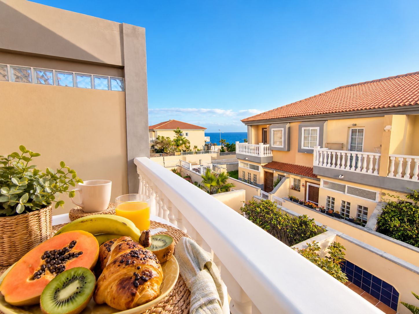 Un balcón con alimentos para el desayuno, vistas a un pueblo costero y un cielo despejado.