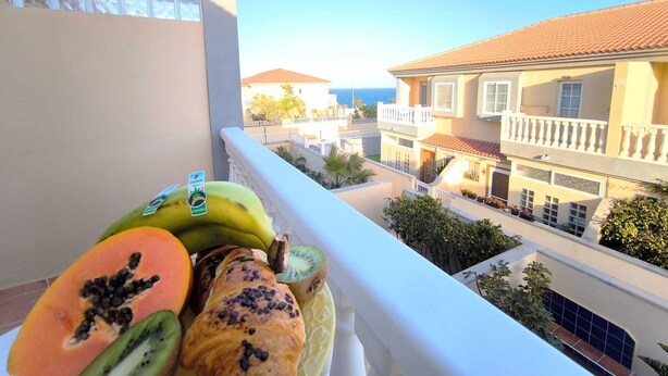 Un balcon donnant sur une ville côtière, une table avec des fruits et une pâtisserie, et un bâtiment avec un toit en tuiles.