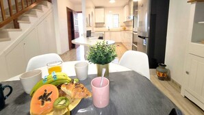 Un coin repas avec une table mise pour le petit-déjeuner, comprenant des fruits et une tasse rose, un escalier et une cuisine en arrière-plan.