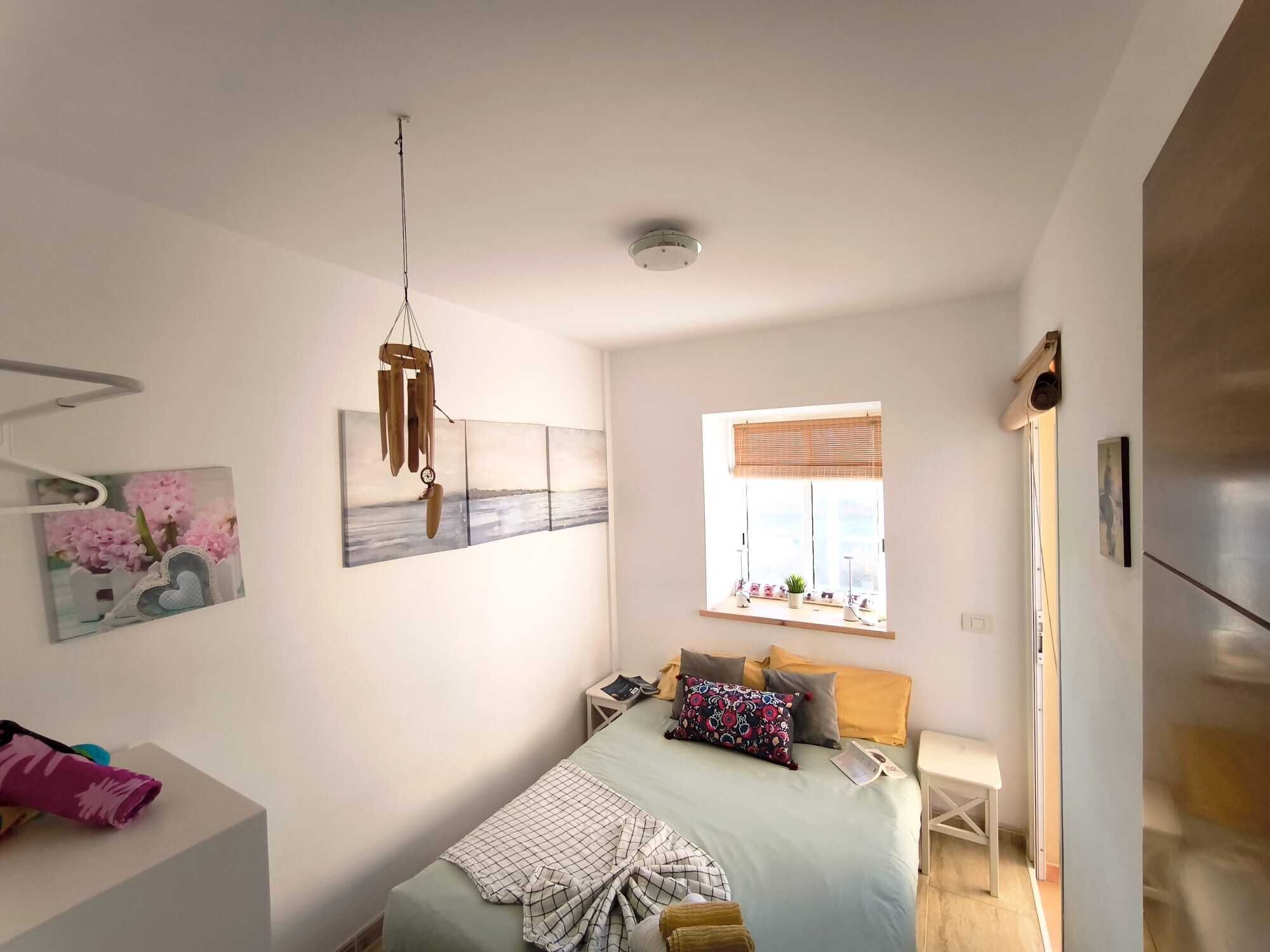 A bedroom with a bed, bedside table, window, and artwork on the wall.