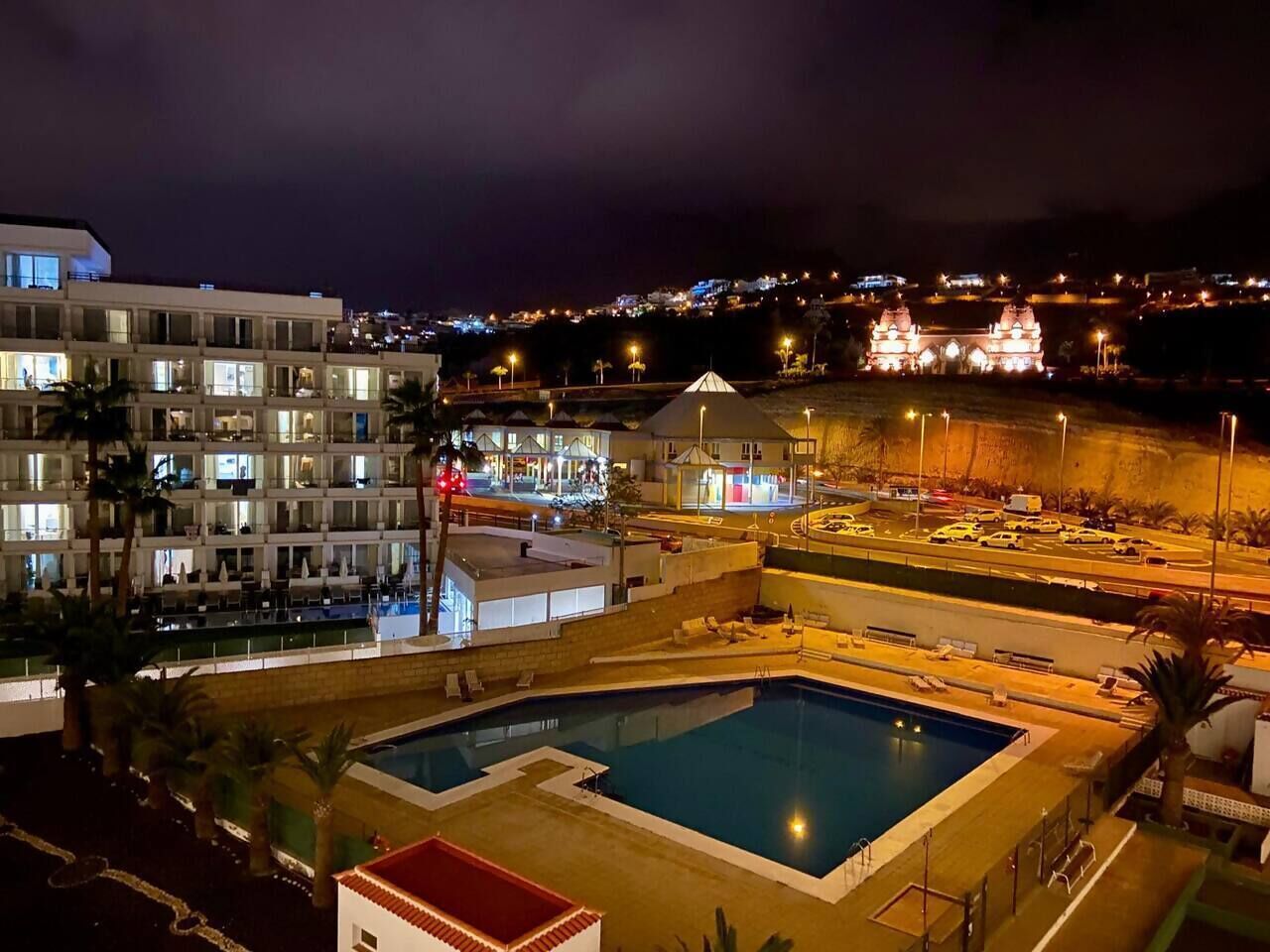 A hotel with a swimming pool, multiple floors, and a view of a hill with a lit-up building.