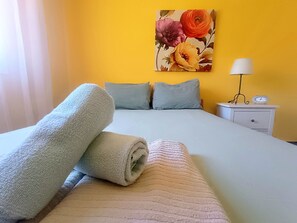 Ein ordentlich gemachtes Bett mit aufgerollten Handtüchern, einer Nachttischlampe und einer Uhr auf einem Nachttisch in einem Zimmer mit gelben Wänden.
