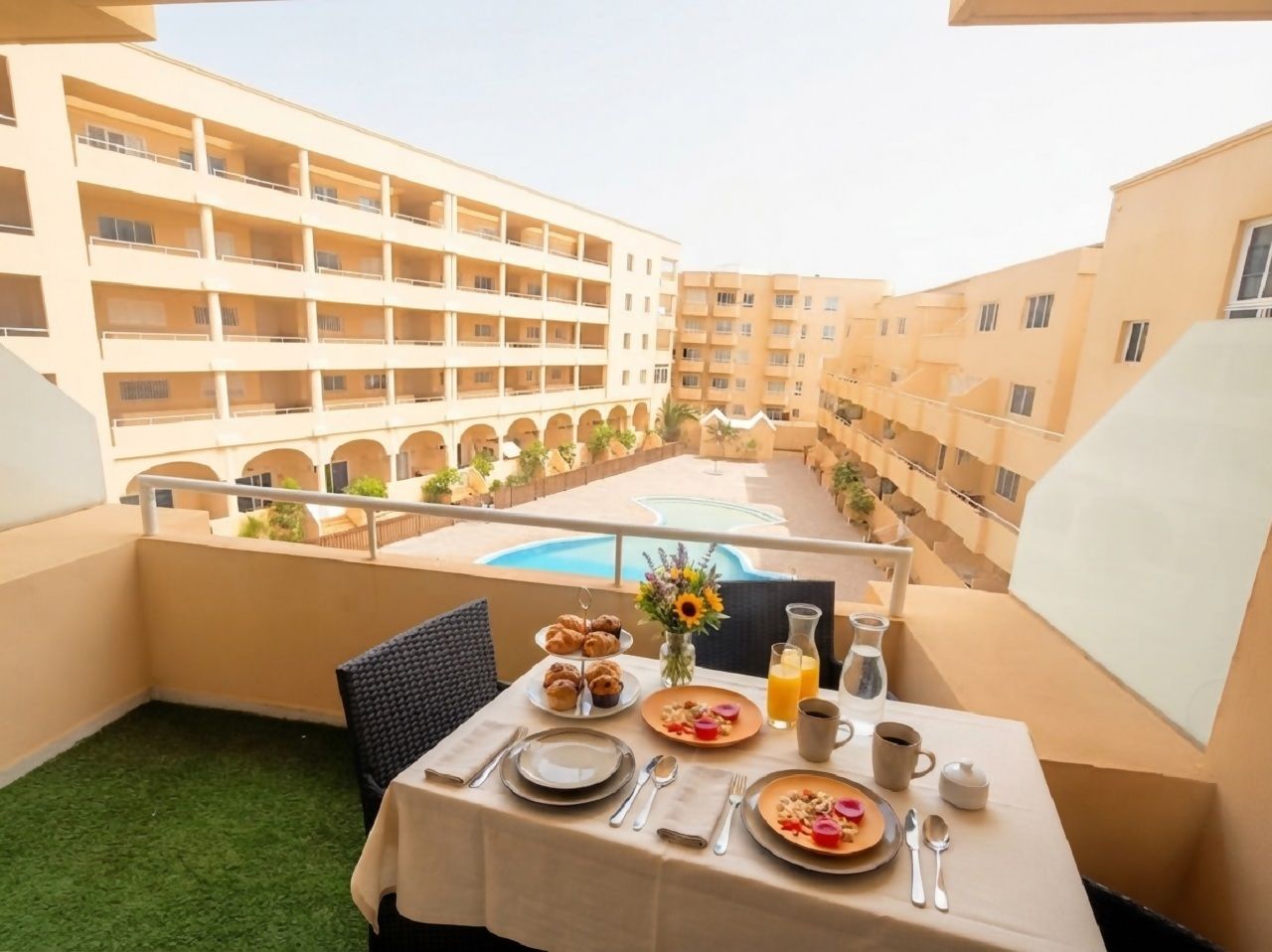 A balcony with a table set for breakfast, overlooking a swimming pool and adjacent buildings.