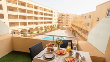 A balcony with a table set for breakfast, overlooking a swimming pool and adjacent buildings.