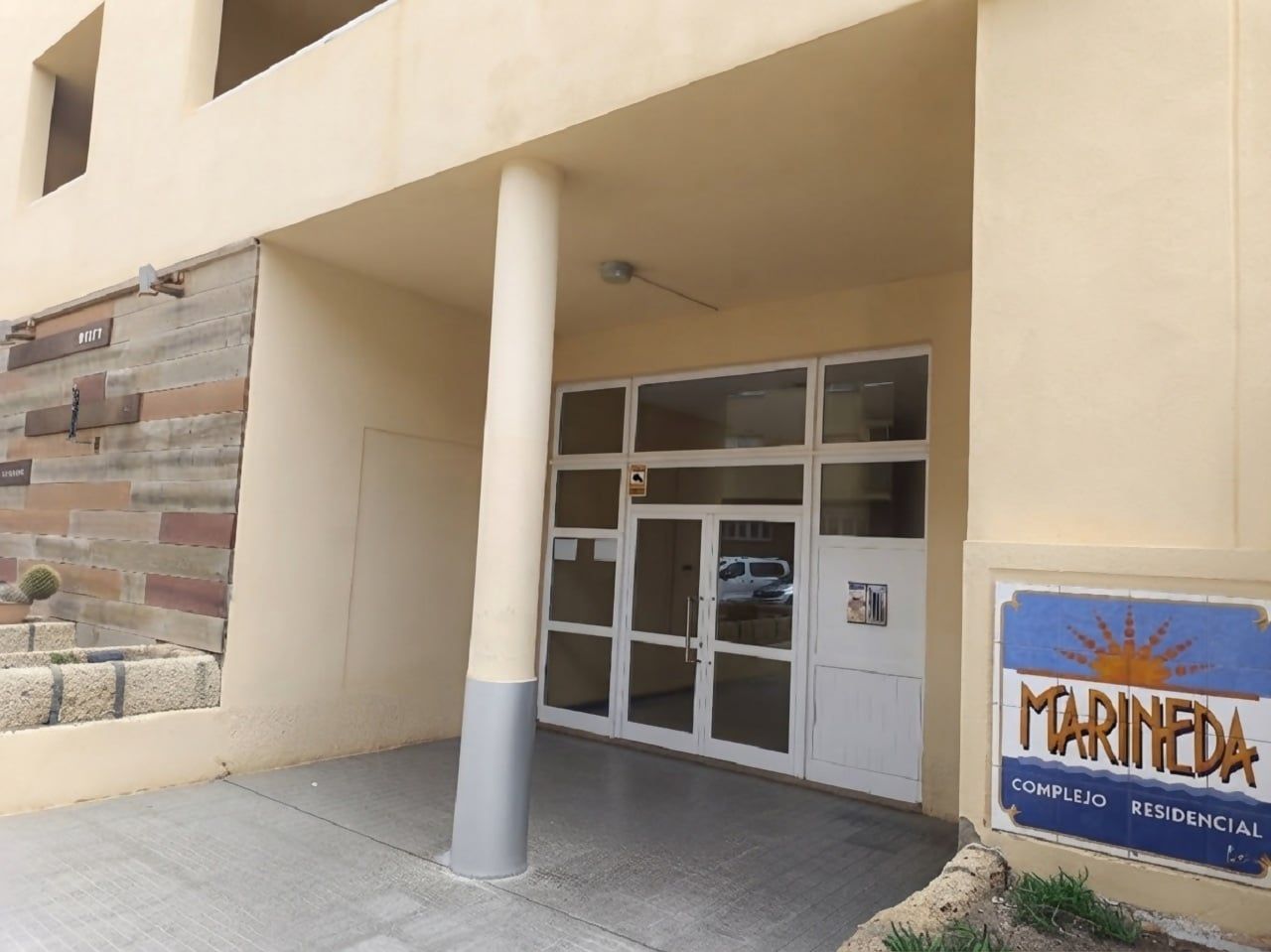 A building entrance with a glass door, a sign reading 'MARINEDA COMPLEJO RESIDENCIAL', and a cactus in a planter.