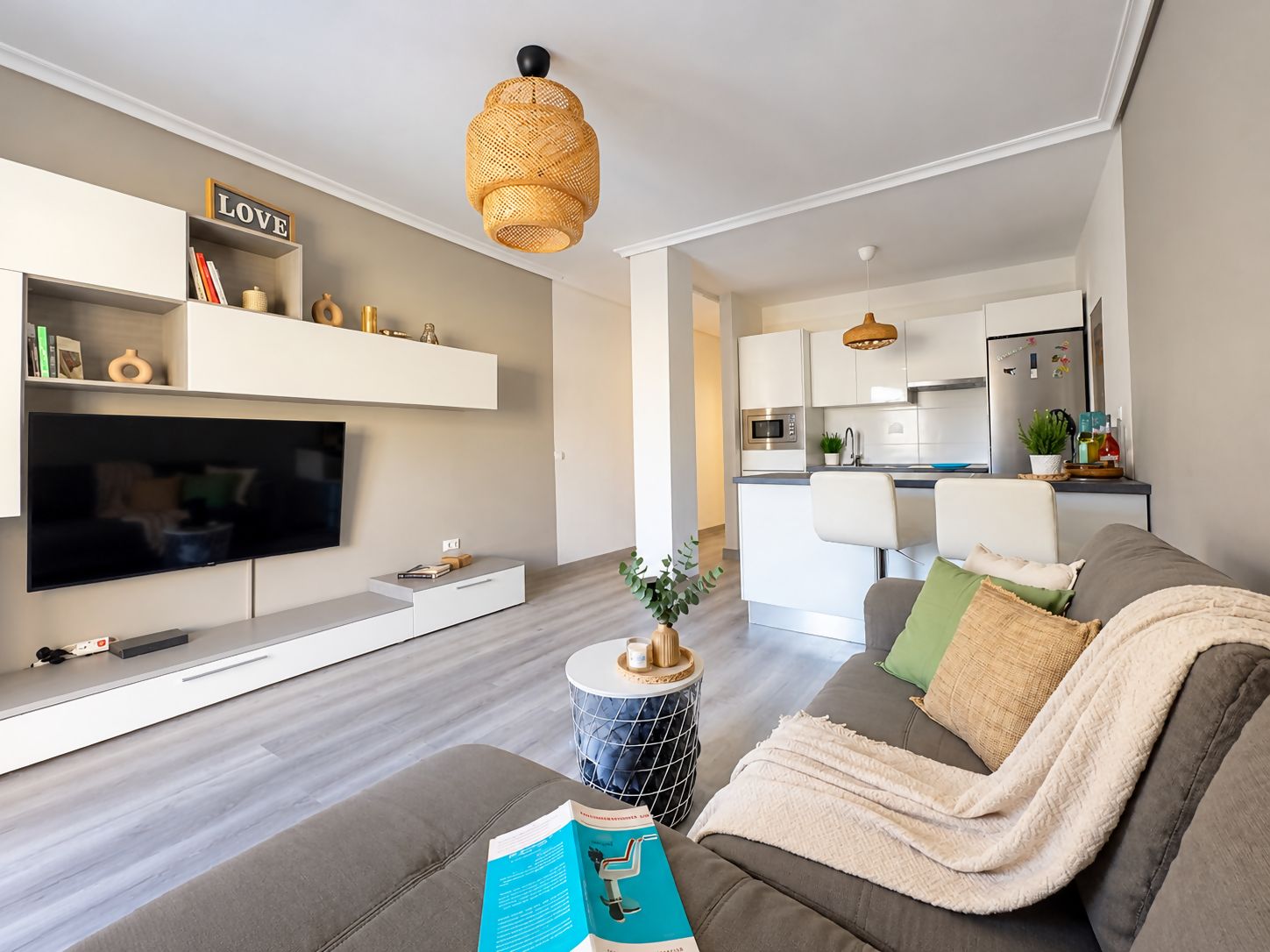 Una sala de estar moderna con un televisor de pantalla plana, un sofá, una mesa de centro con un libro y una lámpara colgante.