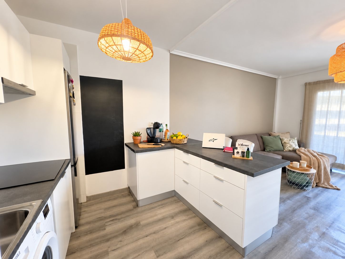 Una cocina compacta con lavadora integrada, cafetera y un pequeño comedor con sofá y una cesta de fruta.