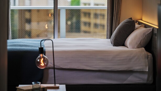 A neatly made bed with pillows, a bedside lamp, and a window with a view of buildings. - Residence Hotel Hakata Sumiyoshi (Fukuoka)