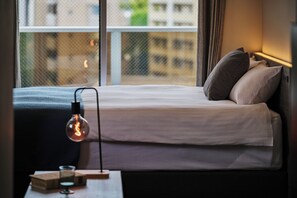 Una cama bien hecha con almohadas, una lámpara de noche y una ventana con vistas a edificios.