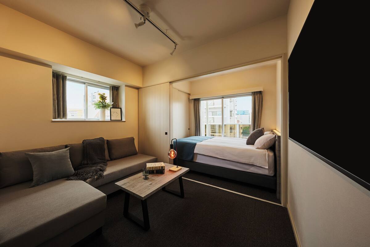 A hotel room with a sofa, a bed, a small table with a lamp, and a view of the city through large windows.