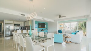 A modern kitchen with a glass dining table, white chairs, and a blue sofa in the living area. - Eagle Beach - 3bed - 3 Bath - Ocean View - Oceania (Oranjestad)