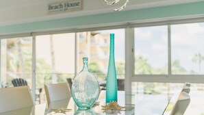 A table with two glass vases and a shell, a sign reading 'Beach House,' and a view of palm trees through large windows. - Eagle Beach - 3bed - 3 Bath - Ocean View - Oceania (Oranjestad)