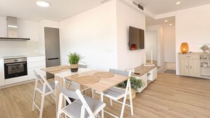 A modern kitchen with white cabinets, a built-in oven, and a dining area with a wooden table and chairs. - 06 Viridis - Golf Terrace - Oliva Nova (Oliva)