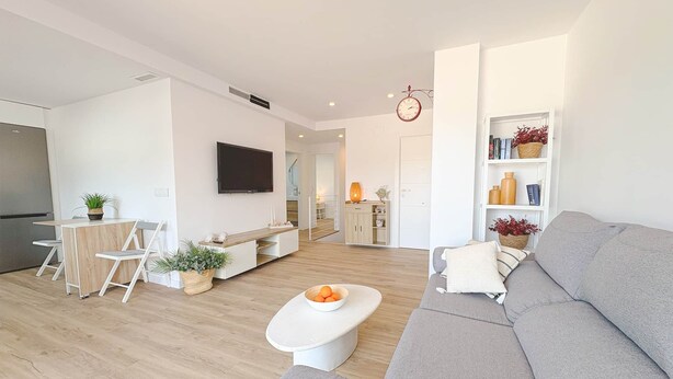 A modern living room with a grey sofa, a white coffee table with fruit, a TV mounted on the wall, and a bookshelf with books and plants. - 06 Viridis - Golf Terrace - Oliva Nova (Oliva)