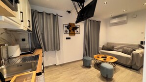 Een moderne woonruimte met een kitchenette, grijze gordijnen, een houten eettafel, een grijze bank en een wandgemonteerde televisie.