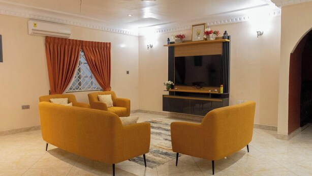A living room with a flat-screen TV, two yellow armchairs, a window with orange curtains, and a fireplace.