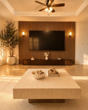 Una sala de estar moderna con un televisor de pantalla plana montado en una pared de paneles de madera, un ventilador de techo, una mesa de centro con objetos decorativos y una planta en maceta.