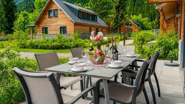Eine Holzhütte mit Essbereich für sechs Personen, umgeben von üppiger Vegetation und einer Bergkulisse.