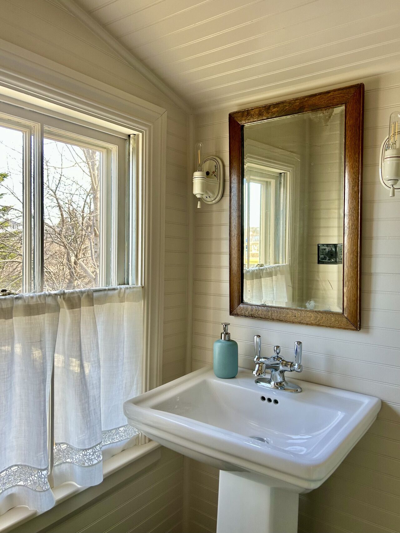 Une salle de bain avec un lavabo blanc, un miroir avec un cadre en bois, un porte-serviettes et une fenêtre avec un rideau.