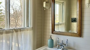 A bathroom with a white sink, a mirror with a wooden frame, a towel rack, and a window with a curtain. - Shackford House in Eastport (Eastport)