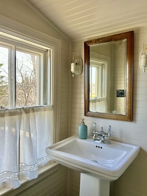 Ein Badezimmer mit einem weißen Waschbecken, einem Spiegel mit Holzrahmen, einem Handtuchhalter und einem Fenster mit Vorhang.