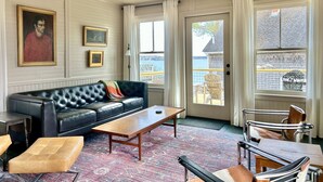A living room with a black leather sofa, a wooden coffee table, a metal chair, and a view of the outside through large windows. - Shackford House in Eastport (Eastport)